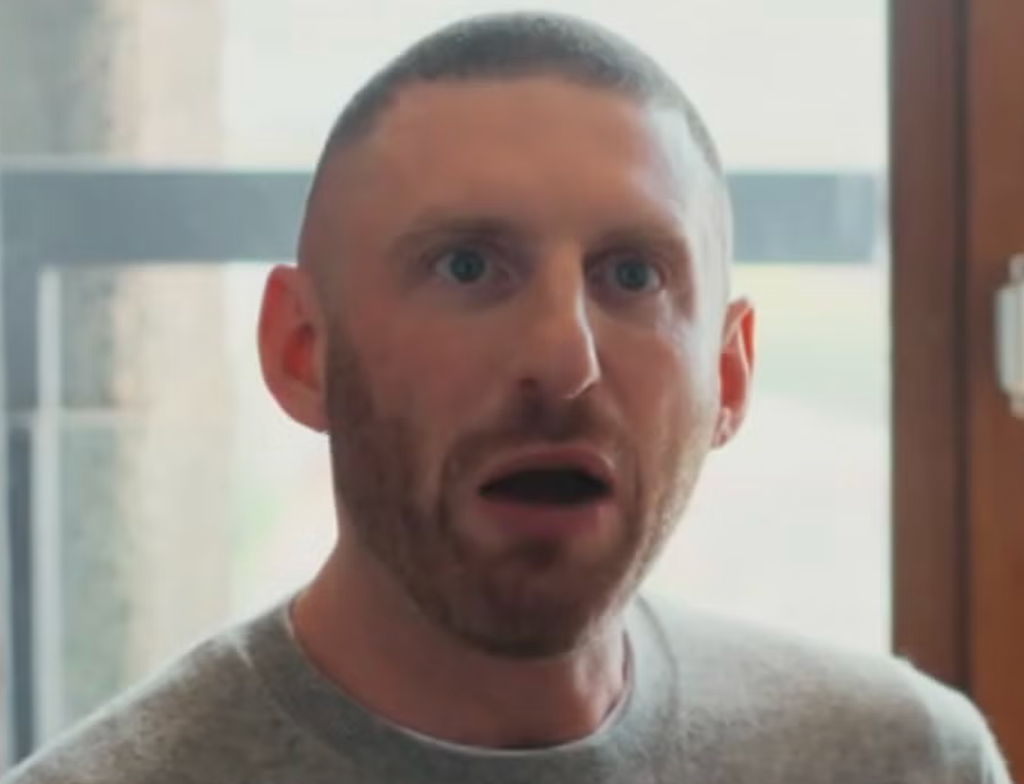 Man with short dark hair and stubble, mouth open in a surprised expression, standing indoors near a window.